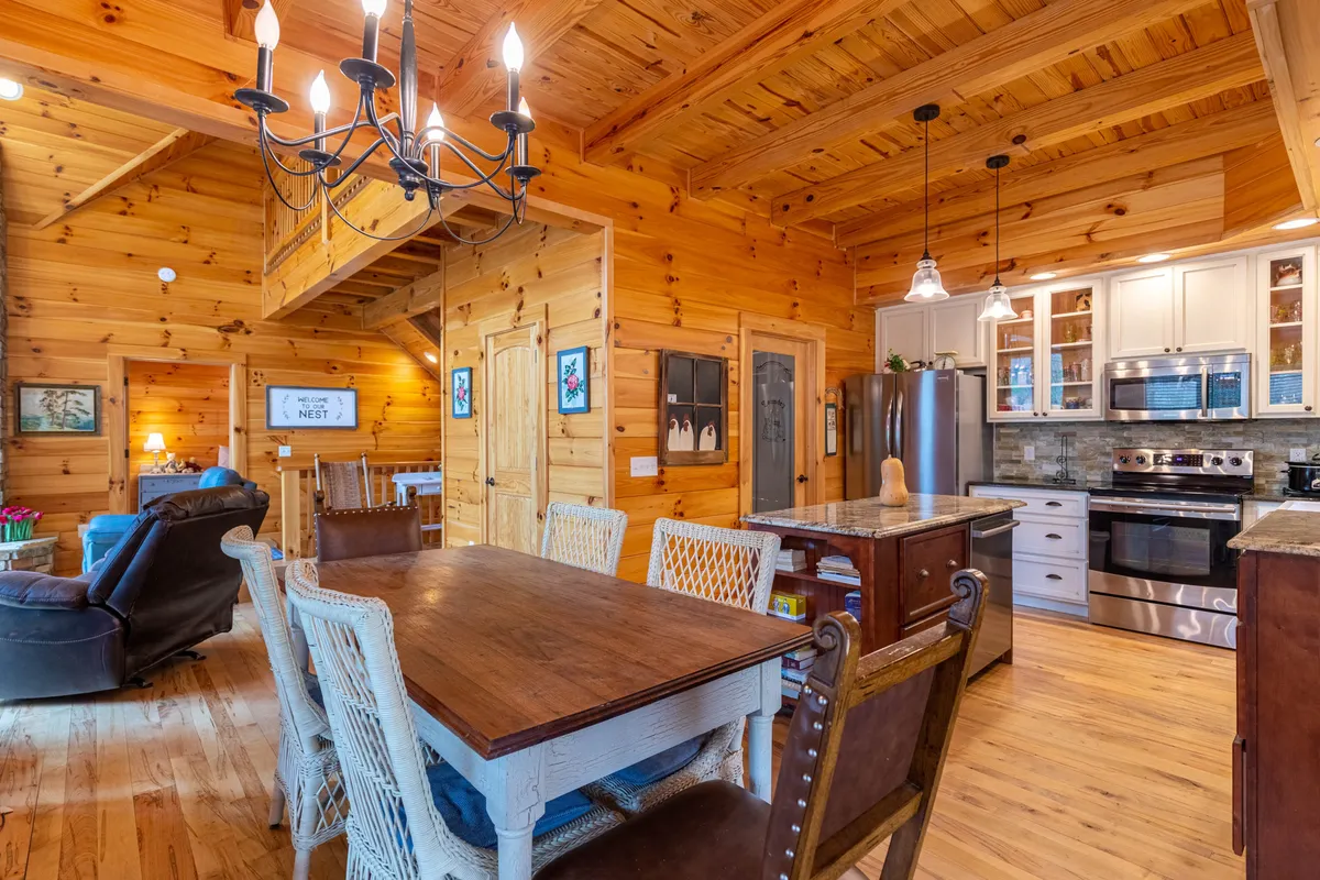 A warm, inviting dining area with a large wooden table under a chandelier, backed by wooden walls and visible kitchen appliances. The space is set for meals, gatherings, and relaxed conversation, reflecting a sense of togetherness.