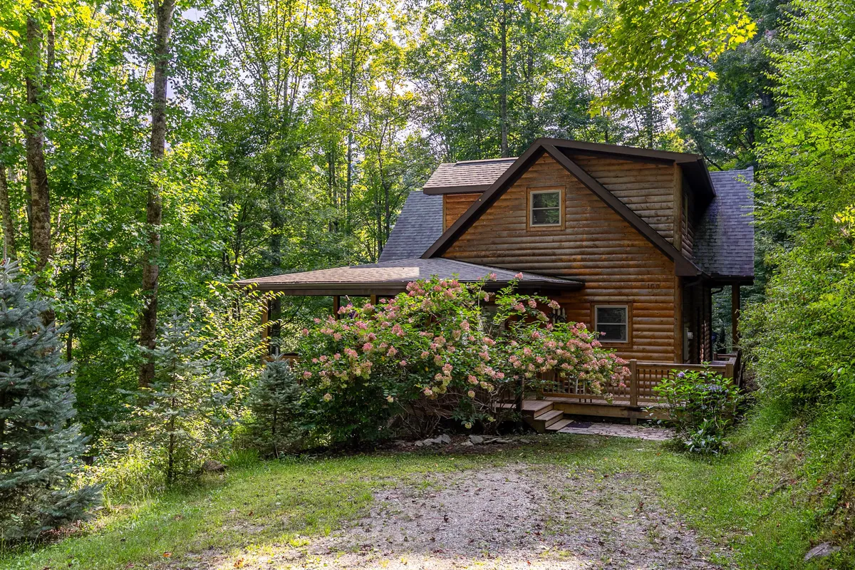 A picturesque exterior view of the log cabin surrounded by flowering bushes and mature trees. The cabin is nestled in nature, with visible greenery and a hint of the peaceful setting that guests can explore, ideal for walks or spotting wildlife.