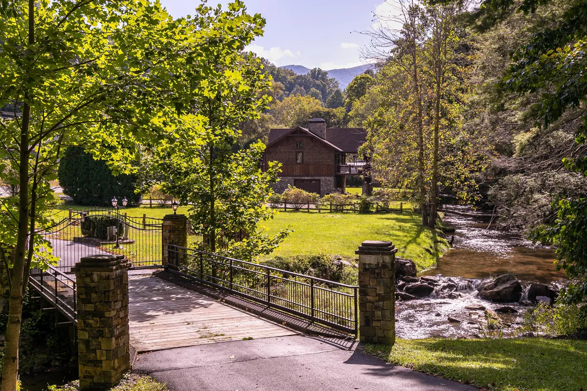 This image captures the cabin from the outside, with the house nestled near a serene stream, a picturesque wooden bridge, and a backdrop of tall trees. The porch, the bridge leading toward the creek, and the lush surroundings all evoke a sense of adventure and relaxation by the water.