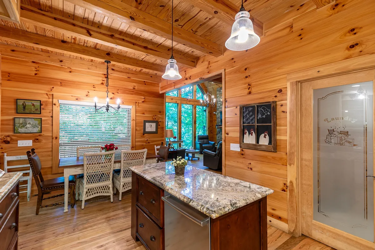 A wide-angle interior shot capturing both the granite kitchen island and the large wooden dining table. Large windows flood the space with natural light and offer a view of surrounding greenery, while decorative light fixtures add a warm glow. The kitchen and dining area are visually connected, emphasizing the open layout.