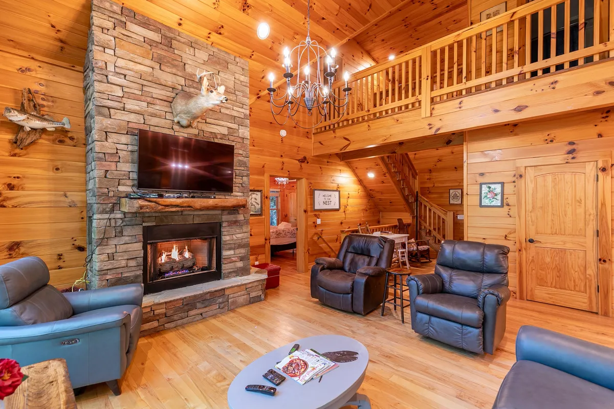 A cozy living room featuring a glowing fireplace, rustic wooden walls, a chandelier, and a comfortable seating area. The space exudes warmth and invites people to gather, with ample room for conversation, relaxation, and watching television together.