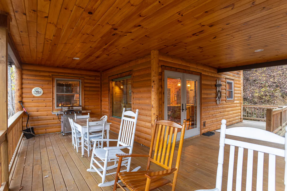 A spacious wooden deck with several rocking chairs, a barbecue grill, and large glass doors leading inside. The area is framed by trees and greenery, suggesting a comfortable space for outdoor dining, relaxing, or gathering after a day of adventure.