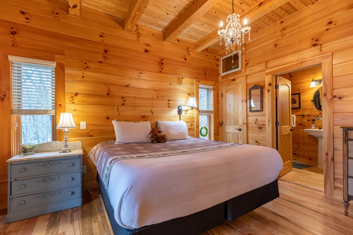 A tranquil bedroom featuring a plush bed with sunlight streaming through large windows, a stylish chandelier overhead, and a cozy nightstand for evening reads. The room feels airy and serene, ideal for unwinding after a day of adventure.