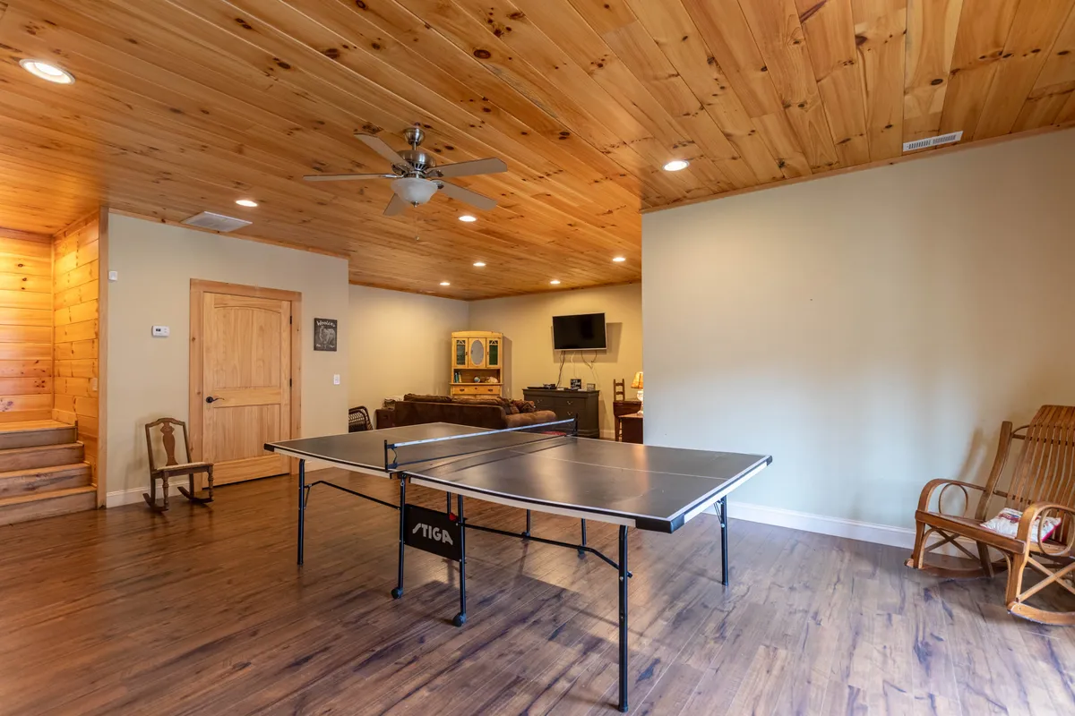 A friendly, casual game room setup with a ping pong table as the focal point, wooden ceiling, and a comfortable seating area nearby. The atmosphere suggests active fun and group interaction, with a backdrop that ties in the cabin's rustic decor.