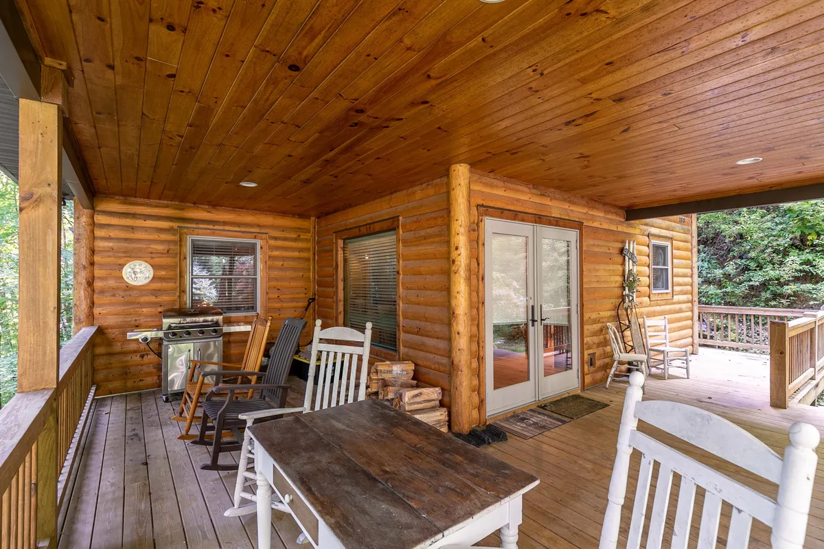 The photo displays a wooden-ceilinged porch with rocking chairs, a dining table, and sliding glass doors opening to the outside. The setting suggests a comfortable transition space between the cabin interior and the outdoors, ideal for relaxing while staying close to nature.
