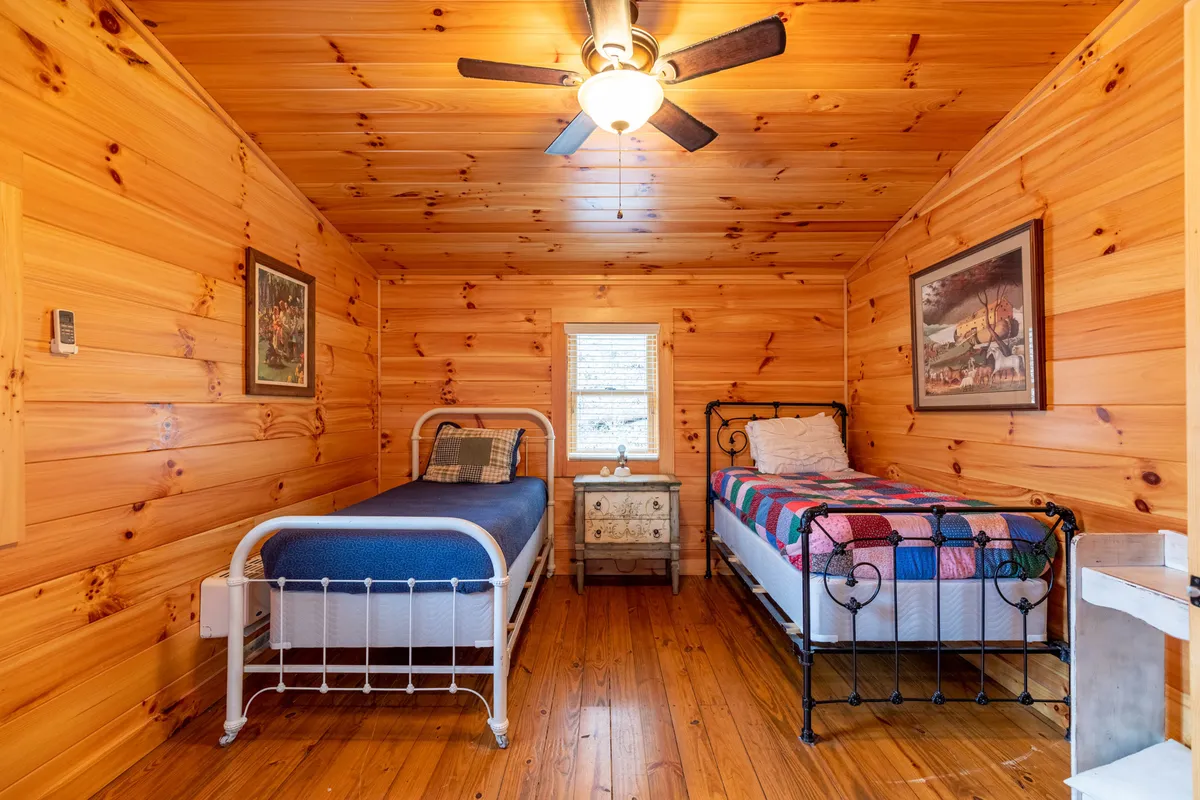 An inviting bedroom with two beds featuring warm wooden wall paneling, a ceiling fan, and a window letting in natural light. The setup is perfect for families or children, suggesting comfort and flexibility.