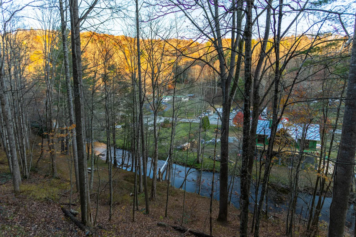 A scenic landscape showing a winding stream bordered by trees, distant mountains on the horizon, and several rustic cabins. The peaceful view highlights the natural beauty and outdoor possibilities of the area, inviting exploration and adventure.