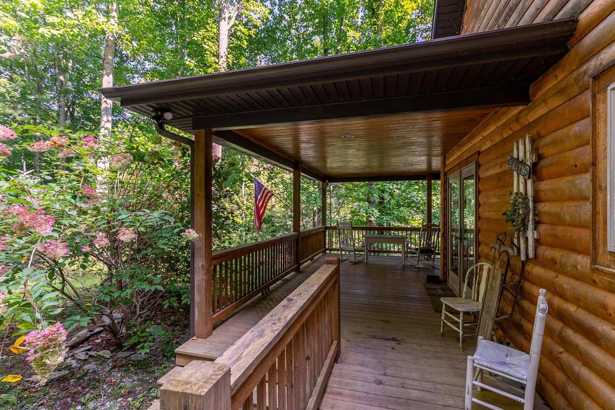 The image shows a charming wooden porch adorned with a flag and colorful flowers. There is a table and chairs set up, suggesting a welcoming spot to relax and enjoy the outdoors. The porch looks out onto greenery, emphasizing its natural surroundings.